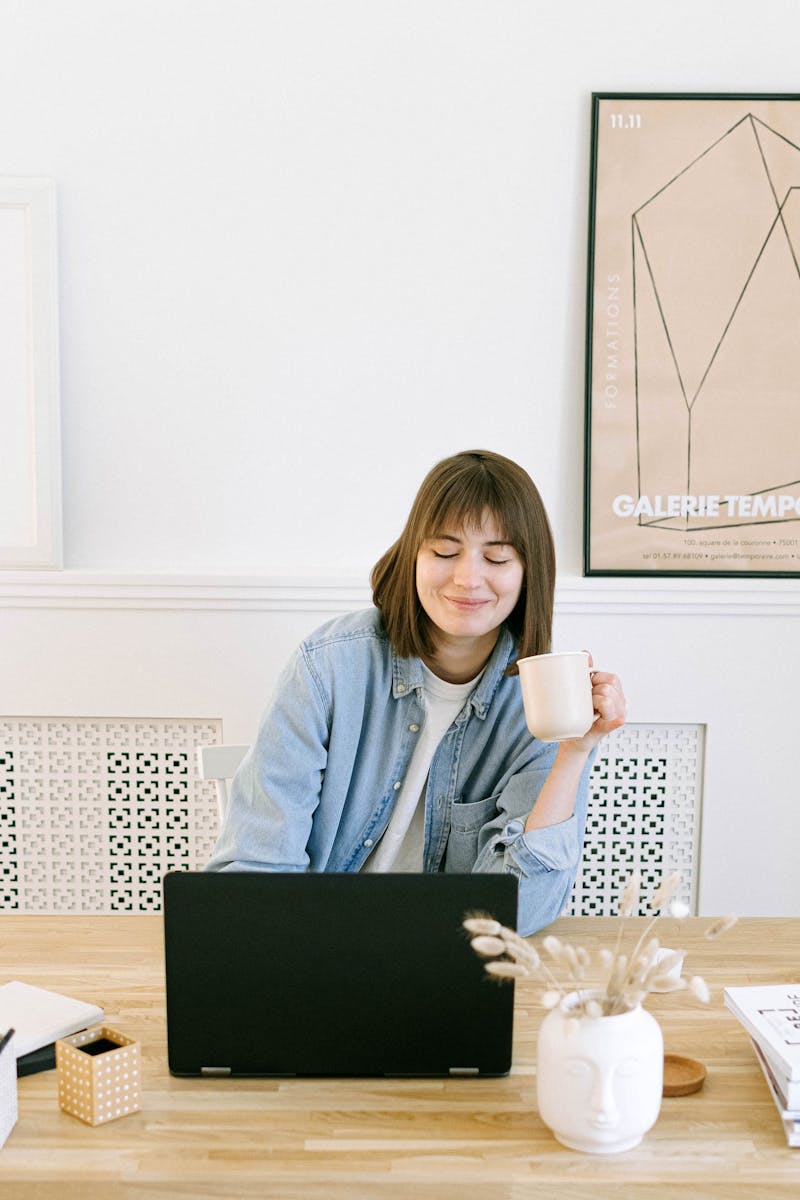 Woman in a home office setting, smiling with a coffee cup near a laptop.